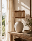 Beige ceramic lamp with woven shade on a wooden table near a window with curtains.