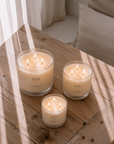 Three SOL Camomile scented candles in clear glass jars with four wicks each, arranged on a rustic wooden table in soft, dappled sunlight.