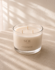 A three-wick soy candle in a clear glass jar with cream-colored wax, labeled 'SOL CASHMERE', set on a textured beige surface with soft window shadow patterns.