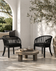 Two black wicker chairs with a wooden coffee table on a patio with a white wall and greenery.