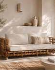 Wicker sofa with beige cushions in a sunlit room with plants and decor.
