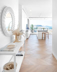 A bright and airy coastal home featuring herringbone oak flooring, a sculptural white mirror, and Kulala Living solid oak bar stools at a marble island. Floor-to-ceiling glass doors open to an ocean-view balcony, blending indoor and outdoor living seamlessly