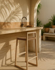 Adanna Bar Chair with oak frame and natural woven seat styled in a Mediterranean-inspired outdoor kitchen setting with tiled benchtop and curved architectural details.