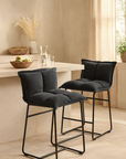 Charcoal padded bar stools with black metal frames styled at a light stone breakfast bar, set in a soft neutral kitchen space with natural decor and warm filtered light.
