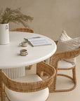 Coastal-inspired dining space featuring curved rattan dining chairs with upholstered seats, paired with a white fluted dining table. Styled with a ceramic vase, neutral-toned decor, organic ambiance