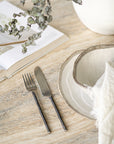 A minimalist table setting featuring hand-forged stainless steel cutlery with sleek black handles, paired with rustic ceramic dishware on a textured stone table, evoking a refined yet organic dining experience.