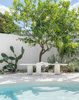KULALA Living Ayanda Concrete Bench in Eggshell styled with the Fara Linen Bath Towel and Small Throw beside a swimming pool, surrounded by a leafy citrus tree, cacti and lush coastal garden greenery.