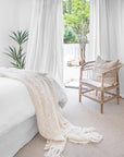 A serene bedroom featuring a neutral-toned upholstered bed with soft layered bedding, a natural rattan armchair with textured cushion for a calming retreat.