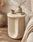 Berber side table in driftstone finish with sculptural curved form styled beside a bed in a soft neutral bedroom setting.