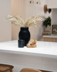 A modern kitchen countertop featuring a striking black ceramic vase with dried foliage, a black Kulala Living candle, and a wooden coaster set. The scene is complemented by warm wood tones and a neutral palette, creating a refined and inviting aesthetic