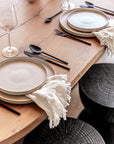 A refined dining table setting featuring KULALA Living’s matte black cutlery, layered ceramic plates, and soft linen napkins on a beautifully crafted timber table. The elegant tableware is complemented by black timber stools, creating a sophisticated and contemporary dining aesthetic
