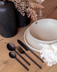 A contemporary dining table setting featuring KULALA Living’s sleek black cutlery, speckled ceramic plates, and a soft linen napkin on a warm timber table. Styled with black sculptural vases and delicate dried foliage, this setting embodies modern elegance and refined simplicity