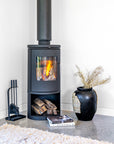 Black decorative vase with natural woven detail styled beside a lit wood fireplace on a polished concrete floor with books and fireplace tools