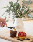 A delicate dessert moment featuring KULALA Living’s black hand-forged butter knife, elegantly spreading cream onto a caramelised fig and nut pastry. Paired with a fresh strawberry tart and displayed on a natural timber board, this scene evokes refined indulgence and artisanal charm