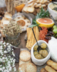 A beautifully styled grazing table featuring KULALA Living’s black hand-forged olive fork resting in a ceramic bowl of marinated olives. Surrounded by crisp crackers, artisanal bread, gourmet cheese, and fresh citrus, this setting embodies effortless entertaining with refined, rustic charm