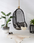A modern black hanging chair with an open-weave design, suspended by a rope, styled with a monochrome cushion and a faux fur throw against a white tiled wall with lush greenery
