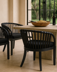 Outdoor dining scene with six black rattan dining chairs around a stone table, styled with a large timber salad bowl and wine, set beside a pool and lush garden backdrop