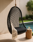 Black hanging outdoor chair with woven frame and cushion, styled with a neutral throw beside a poolside garden setting