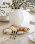 A stylish black mini cheese cutlery set from KULALA Living, featuring a fork, knife, and spreader elegantly arranged on a ceramic platter with brie cheese and crackers, creating a sophisticated and inviting dining scene