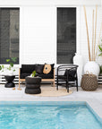 A stylish outdoor lounge setting featuring a black cushioned sofa with a natural wood frame, a black rattan armchair, and textured décor, set beside a serene poolside backdrop.