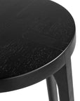A sleek black oak round stool featuring a minimalist design with subtly curved legs. Crafted from solid oak with a matte black finish, this versatile piece functions as a stool or side table, adding a modern touch to any space