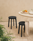 Pair of black outdoor stools with round seats styled in a sunlit courtyard next to a travertine table set with summer fruit and glassware