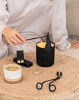 KULALA Living black soy candle being snuffed out, styled with candle care tools, ceramic coffee cup, and matchboxes on a woven table