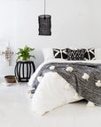 Black wooden side table with open slat design styled in a contemporary bedroom with monochrome accents, featuring textured cushions, a woven pendant light, and pom-pom throw.