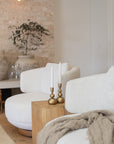 A beautifully styled corner featuring a white boucle swivel chair paired with a natural timber side table. Brass candlesticks with white taper candles add warmth, while a soft textured throw drapes over the chair. A rustic stone vase with greenery sits against an exposed brick wall, creating an inviting and serene living space.