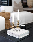 A refined coffee table vignette featuring KULALA Living’s brass candlestick holders with elegant tapered candles, paired with design books and a black minimalist candle. The setting is styled against a white linen sofa, a sculptural wooden side table, and neutral-toned accents, embodying a serene and sophisticated living space