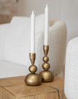 A pair of handcrafted brass candlesticks with white taper candles, elegantly styled on a solid timber side table. The warm brass tones contrast beautifully against the soft boucle chairs in the background, adding a touch of sophistication and warmth to the space