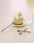 A luxurious dessert setting featuring KULALA Living brass dessert spoons, a matcha cheesecake topped with macarons, and a citrus-infused cocktail. A hand is seen delicately scooping a bite, embodying the essence of slow living and refined indulgence