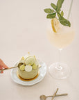A luxurious dessert setting featuring KULALA Living brass dessert spoons, a matcha cheesecake topped with macarons, and a citrus-infused cocktail. A hand is seen delicately scooping a bite, embodying the essence of slow living and refined indulgence