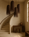 Cluster of KULALA Chiku Pendants in dark natural fibres suspended above a sculptural staircase with soft, earthy-toned walls and timber console