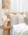 A serene coastal-inspired bedroom featuring a rattan bedhead, layered neutral linen cushions, a natural oak side table, and a woven pendant light, creating a relaxed and airy aesthetic