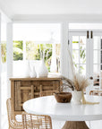 A serene coastal dining space featuring a white round dining table with a sculpted wooden base, surrounded by natural rattan dining chairs. A rustic timber sideboard and white ceramic vases enhance the organic aesthetic. A vase with dried floral arrangements adds a soft, natural touch