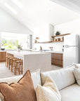 Open-plan coastal kitchen and lounge featuring a neutral-toned sofa, natural timber bar stools, stone island, and a white Smeg fridge under vaulted ceilings.