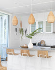 Bright coastal kitchen with natural rattan pendant lights, wooden and metal bar stools, white cabinetry, and a neutral-toned stone benchtop, creating a fresh and inviting space with organic textures