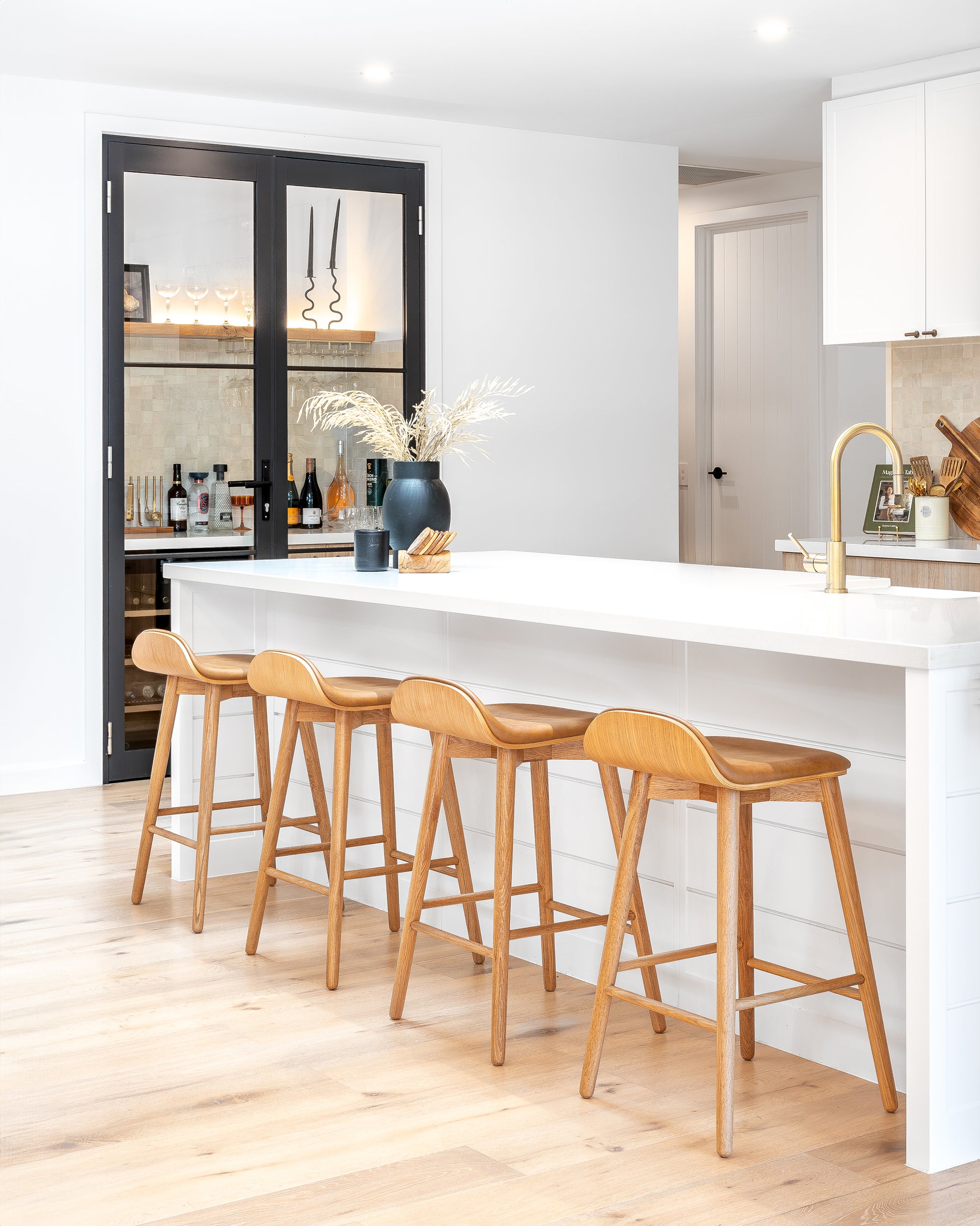 A modern coastal kitchen featuring a sleek white island with four wooden bar stools, a gold kitchen tap, and a stylish home bar with glass shelving and a black-framed cabinet, creating a sophisticated yet inviting atmosphere