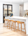A modern coastal kitchen featuring a sleek white island with four wooden bar stools, a gold kitchen tap, and a stylish home bar with glass shelving and a black-framed cabinet, creating a sophisticated yet inviting atmosphere