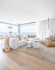 Coastal-inspired living room featuring a white modular sofa with textured cushions, a woven jute pouf, and a suspended white rattan hanging chair by a floor-to-ceiling window with ocean views.