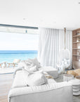Coastal-inspired living room featuring a white modular sofa with textured cushions, a woven jute pouf, and a suspended white rattan hanging chair by a floor-to-ceiling window with ocean views
