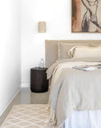 Dark timber side table styled beside a natural linen-draped bed with neutral decor in a softly lit contemporary bedroom