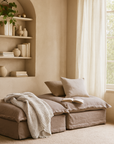 Earth-toned linen daybed with matching cushions, knitted throw, and an open book, styled beside arched shelves with neutral decor and sheer curtains.