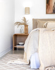 Detail of the Flynn Open Bedside Table in natural oak, styled with a sculptural timber vase, glassware, and neutral-toned bedding in a soft, sunlit bedroom