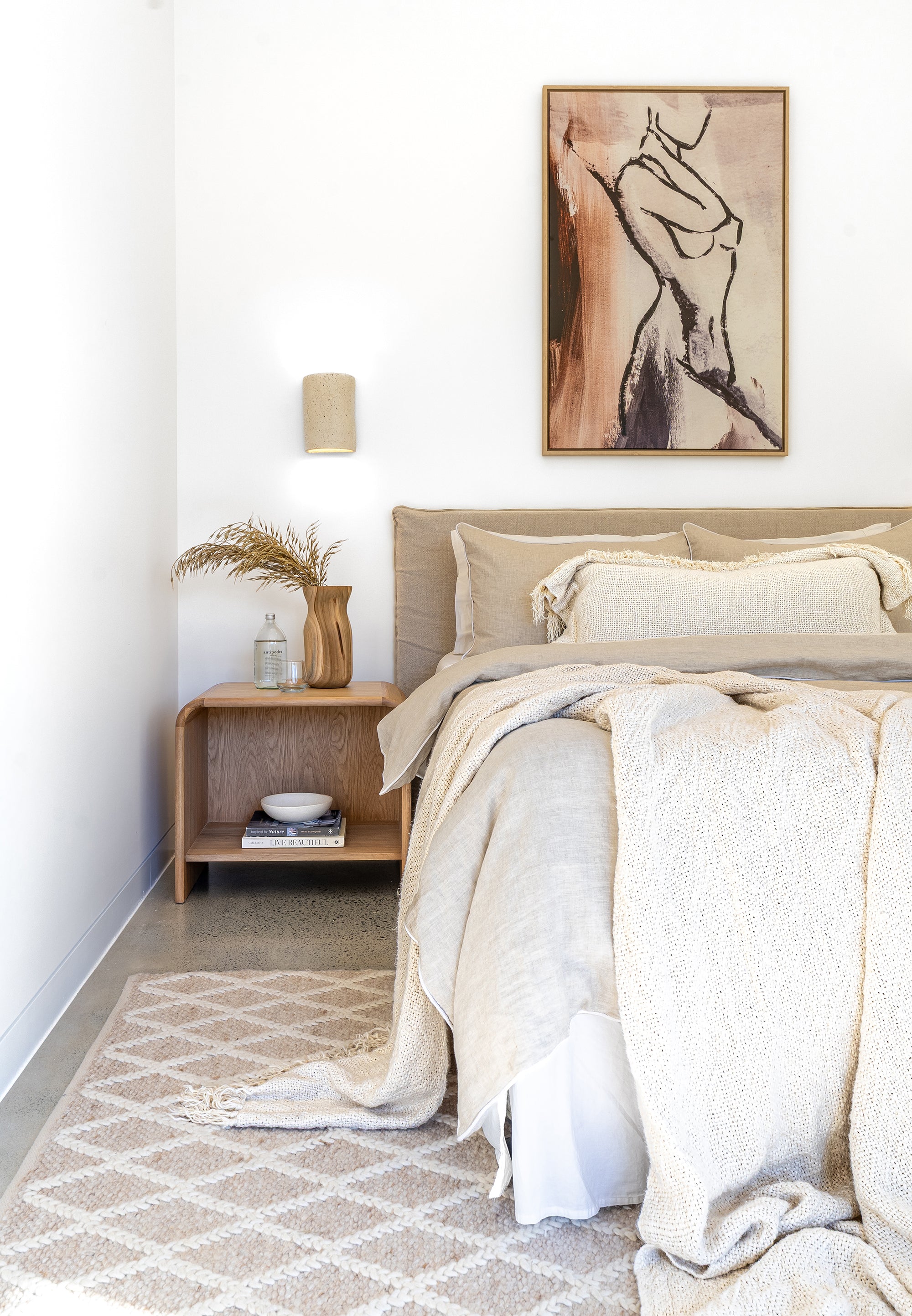 Neutral-toned bedroom featuring the Flynn Open Bedside Table in natural oak, styled with sculptural decor, layered linen bedding, and a textured rug beneath a contemporary artwork