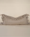 A natural-toned lumbar cushion with a woven texture, featuring delicate fringed edges, set against a neutral backdrop