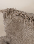 A natural-toned lumbar cushion with a woven texture, featuring delicate fringed edges, set against a neutral backdrop