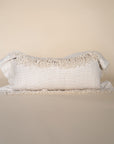A natural-toned lumbar cushion with a woven texture, featuring delicate fringed edges, set against a neutral backdrop