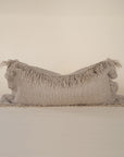 A natural-toned lumbar cushion with a woven texture, featuring delicate fringed edges, set against a neutral backdrop